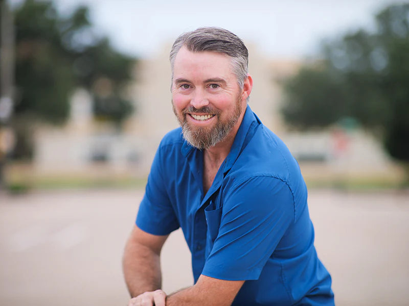 Joel Higgins in a blue shirt sitting outdoors with trees in the background