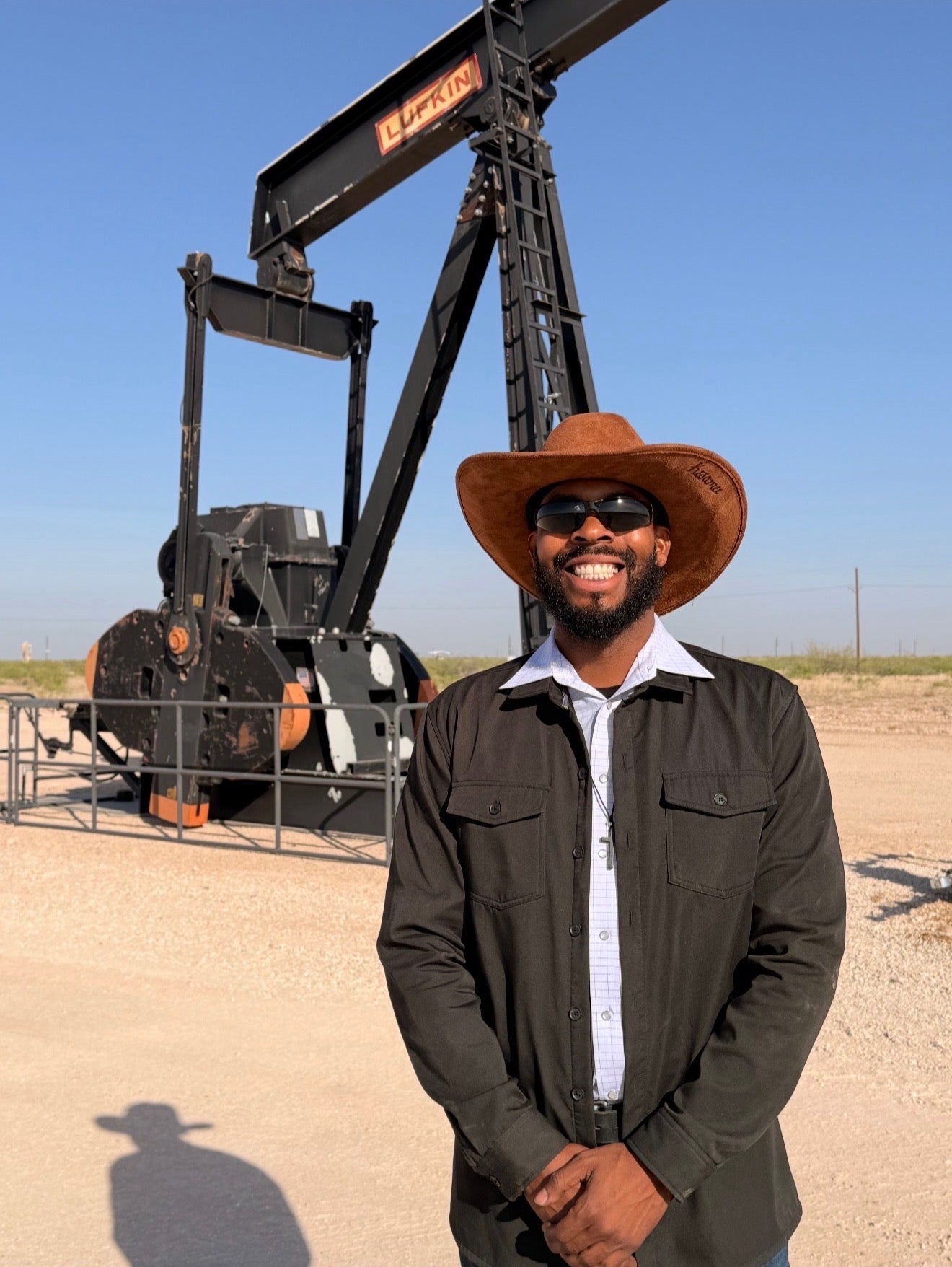 Man wearing a cowboy hat standing in front of an oil pumpjack.