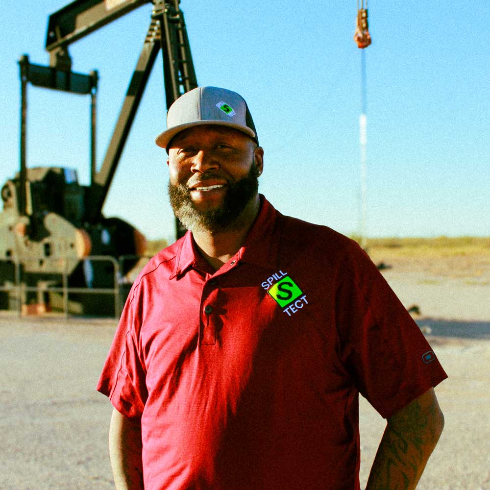 Tyari Laster standing outdoors in a red shirt and grey hat, in front of an oil derrick pump